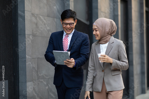 Young business muslim woman and business man colleague walking and discussing outside the office. Woman is holding a coffee and businessman is holding a digital tablet outdoor.