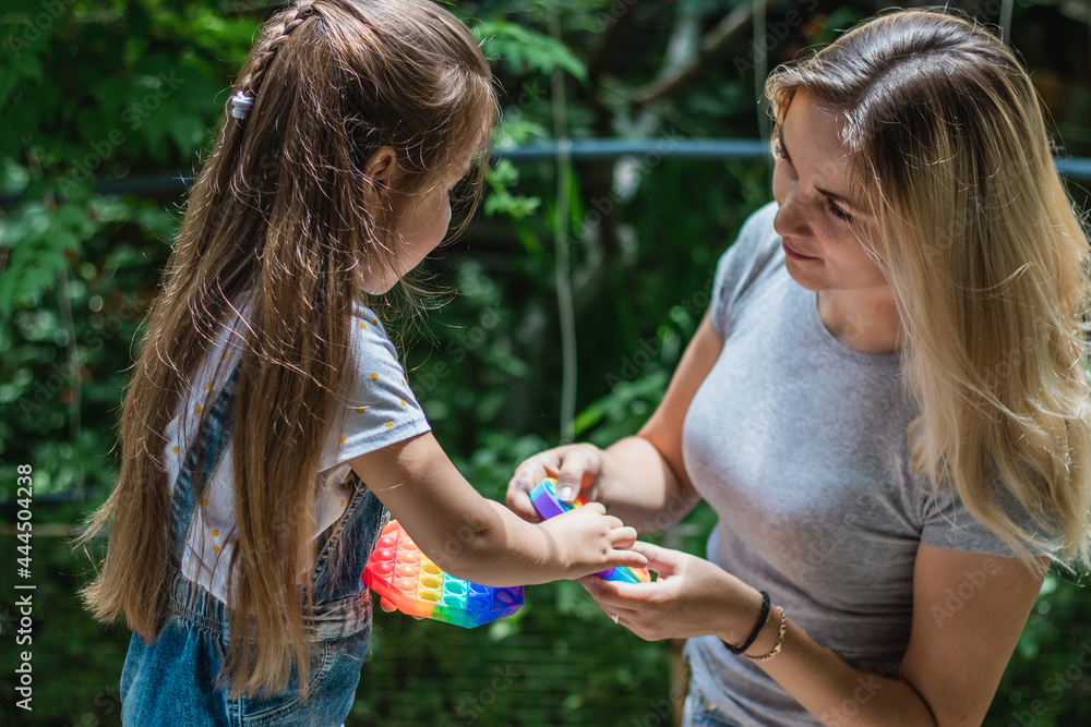 Naklejka premium A child and his mother playing with a pop-fidget on a green background. A girl and a mother click on a colored rainbow silicone anti-stress toy Push pop bubble. Communication with children.