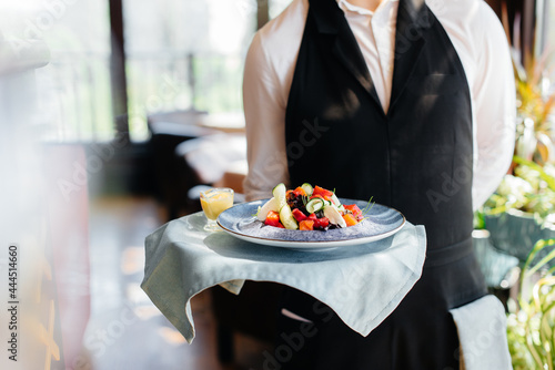 A young waiter in a stylish uniform stands with an exquisite dish on a tray near the table in a beautiful restaurant close-up. Restaurant activity, of the highest level.