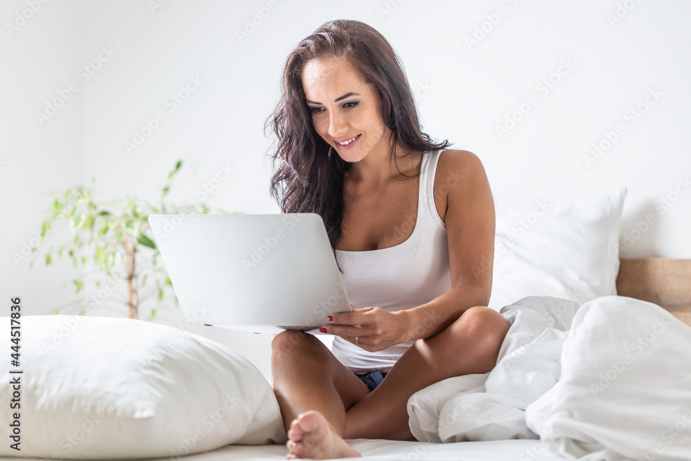 Woman works on her laptop sitting on a bed surrounded by white pillows Woman works on her laptop sitting on a bed surrounded by white pillows