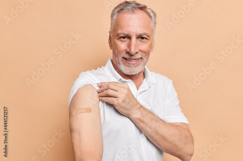 Fototapete Old bearded man got vaccinated against coronavirus shows arms with adhesive plaster cares about healthy during pandemic poses against brown background