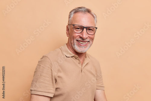 Happy grandpa senior man with grey hair and beard smiles toothily shows perfect white teeth wears optical glasses and casual t shirt enjoys life glad to meed old friend isolated over beige background photo