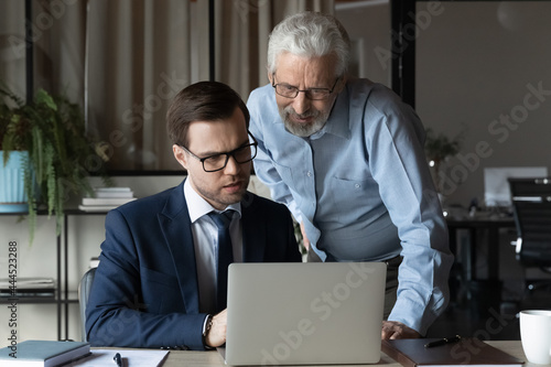 Older professional giving help, advice to young coworker, explaining issues, supervising work of new employee. Businessmen discussing project at laptop, looking at screen. Mentor training intern