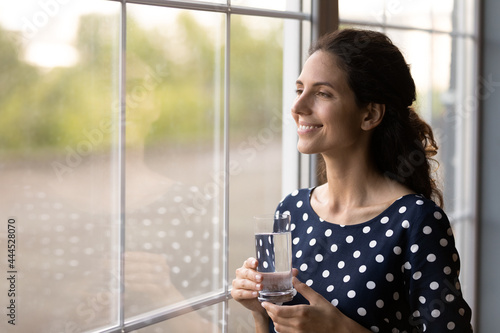 Happy Latin woman in casual holding glass of pure fresh potable water  drinking  keeping healthy metabolism  looking out window  thinking and smiling. Hydration  healthcare concept