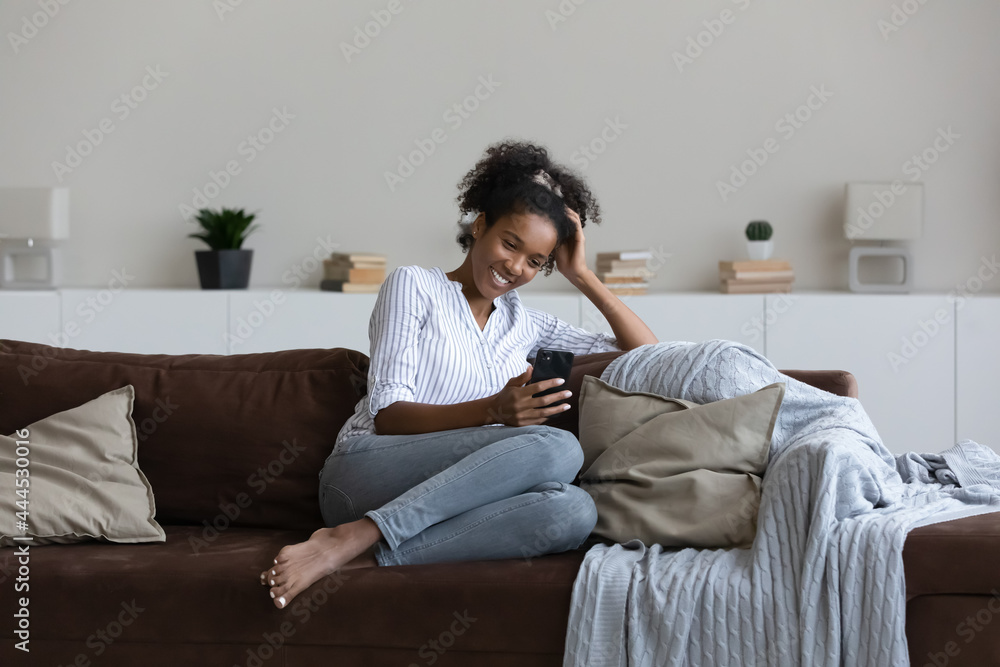 Smiling African American woman using smartphone, sitting on cozy couch at home, excited young female looking at phone screen, chatting in social network online, enjoying leisure time with device Smiling African American woman using smartphone, sitting on cozy couch at home, excited young female looking at phone screen, chatting in social network online, enjoying leisure time with device
