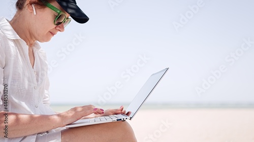 Mature woman using laptot on background the sea and blue sky. Remote work, summer vacation concept, beach work, freelance work and leisure photo