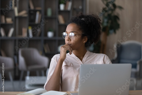 Thoughtful African American woman in glasses looking to aside touching chin, pondering project strategy or ideas, pensive young female student or freelancer sitting at work desk with laptop photo