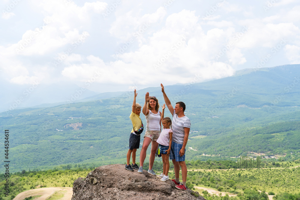 Happy family with children with their hands raised stands on rocks on hill and looks at camera. Concept of family trip in mountains. Father, mother, daughter and son have fun in nature