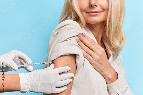 Nice caucasian blond unrecognizable lady gets her vaccine and smiles, comes to vaccination centre in a dress. Nurse injects carefully the vaccine, wears white gloves. Isolated over blue background. photo