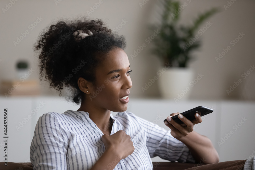 Close up smiling African American recording audio message on smartphone, sitting on couch at home, happy young female chatting with friends in social networks, making phone call by speakerphone Close up smiling African American recording audio message on smartphone, sitting on couch at home, happy young female chatting with friends in social networks, making phone call by speakerphone