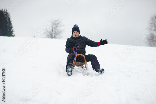 adult man in winter black clothes enjoys a ride on a wooden sledge with a smile and tries not to fall off it. Foot braking. Use of fresh powder for a fast ride. Winter season in the Czech Republic