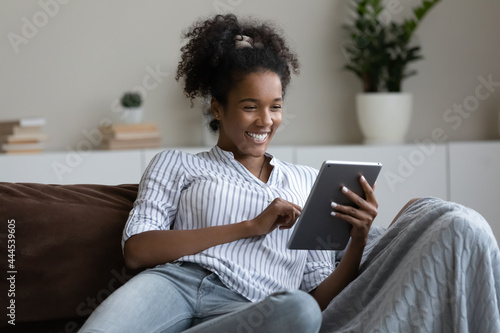 Excited African American woman having fun with tablet, sitting on couch at home, happy positive young female looking at device screen, chatting in social network, laughing at funny joke, playing game