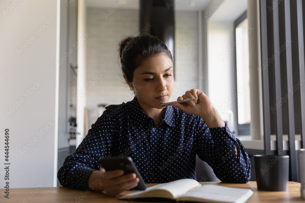 Indian millennial student girl preparing for exam, watching learning webinar on smartphone, writing notes in study notebook. Female manager working from home, thinking over calls plan at organizer Indian millennial student girl preparing for exam, watching learning webinar on smartphone, writing notes in study notebook. Female manager working from home, thinking over calls plan at organizer