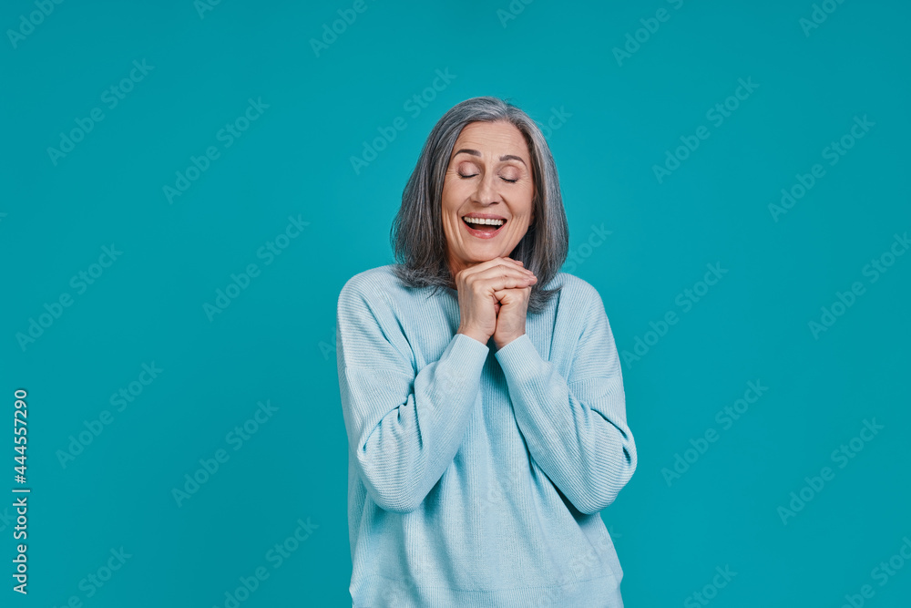 Mature beautiful woman keeping hands clasped and smiling while standing against blue background Mature beautiful woman keeping hands clasped and smiling while standing against blue background