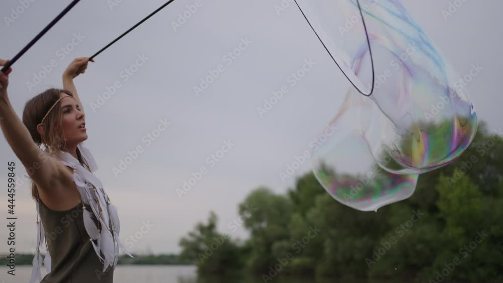A young hippie woman in a dress and with feathers on her head makes huge soap bubbles at sunset on the shore of a lake in slow motion