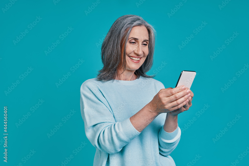 Mature beautiful woman using smart phone and smiling while standing against blue background Mature beautiful woman using smart phone and smiling while standing against blue background