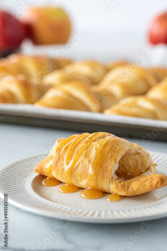 Close up view of an apple crescent roll topped with caramel sauce and a tray of other crescent rolls in the background.