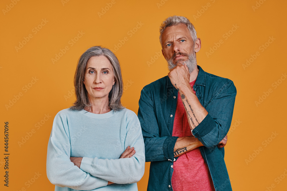 Thoughtful senior couple looking at camera while standing together against orange background Thoughtful senior couple looking at camera while standing together against orange background