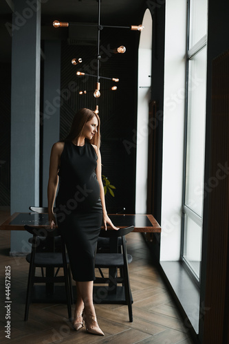 A young pregnant beautiful woman in a black dress is posing in the dark interior of the restaurant and looking aside