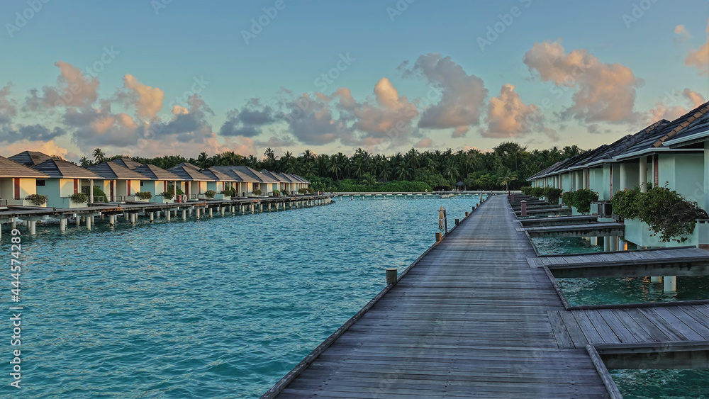 A wooden path leads over the avquamarine ocean to the water villas. In the blue sky, golden morning clouds. Thickets of palm trees on the shore. Maldives