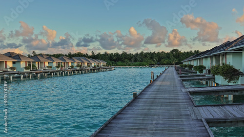Fototapeta Naklejka Na Ścianę i Meble -  A wooden path leads over the avquamarine ocean to the water villas. In the blue sky, golden morning clouds. Thickets of palm trees on the shore. Maldives