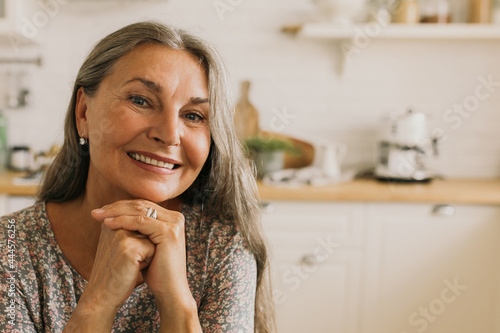 Smiling elderly female sitting at kitchen table. Cheerful middle-aged woman looking at camera with happiness and calmness. Aging and mental balance concept