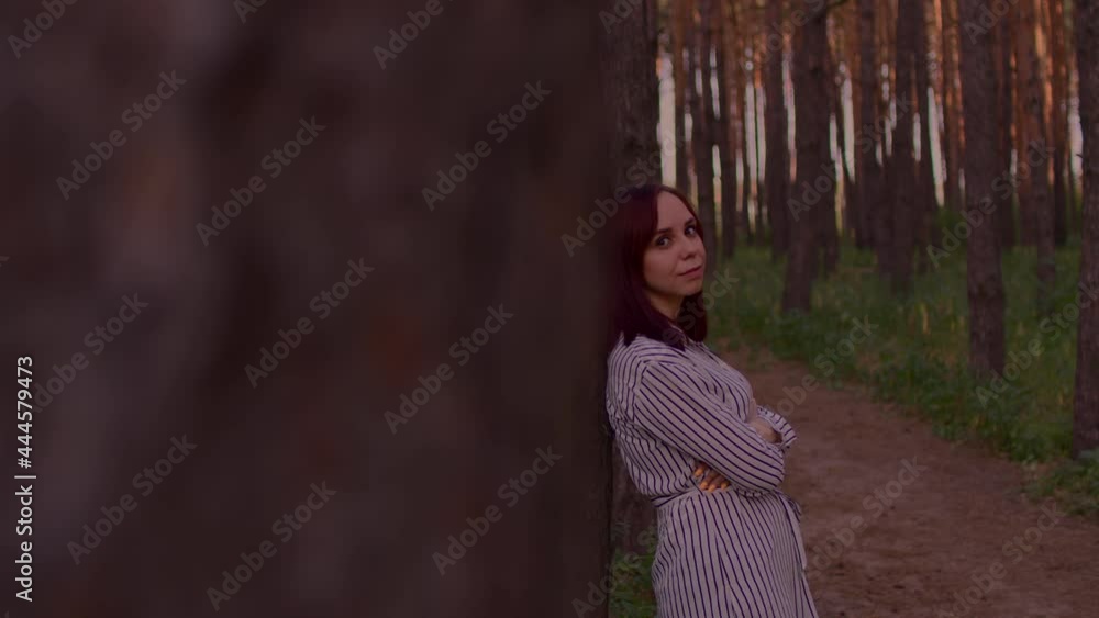Young woman stands, leaning on tree on walk in forest. Camera peeping on pretty female and flirting with her Young woman stands, leaning on tree on walk in forest. Camera peeping on pretty female and flirting with her