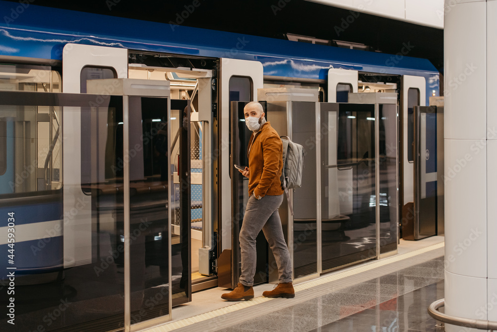 A man in a medical face mask to avoid the spread of coronavirus is holding a smartphone while entering the modern subway car. A bald guy in a surgical mask is keeping social distance.