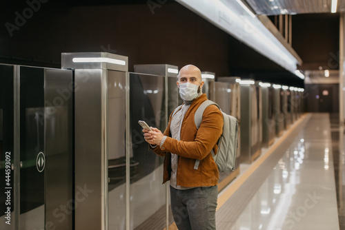 A man in a medical face mask is holding a phone and staring to the side while waiting for a train at the subway platform. A bald guy in a surgical mask is keeping social distance.