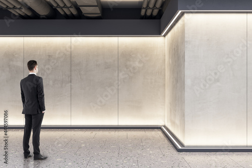 Businessman standing in modern exhibition space concrete interior with mockup place on wall. Mock up.