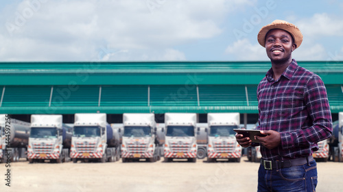 African male truck driver holding tablet and checking route for new destination. In background parked truck vehicles. Concept of transportation service.