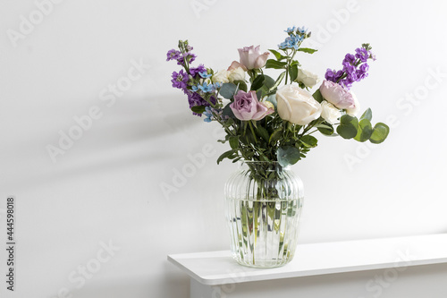 Bouquet of hackelia velutina, purple and white roses, small tea roses, matthiola incana and blue iris in glass vase is on the white coffee table.