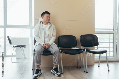 young asian man waiting for an interview or meeting sitting in the hallway in the waiting room. Student or entrant in the reception for exam or employment hr. Male patient in office a hospital clinic