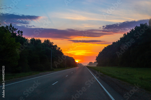 Dawn on the road. The bright sun rises against the background of the sky with clouds, around the forest. Morning landscape