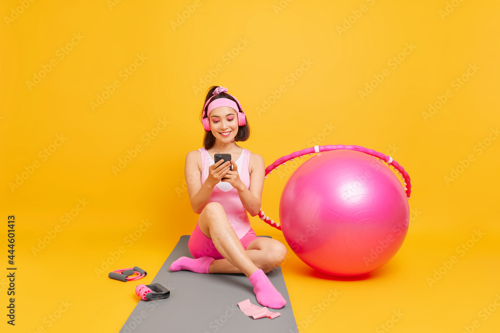 Positive Asian woman with dark hair checks her sport achievements on smartphone application sits on fitness mat dressed in activewear uses swiss ball hula hoop goes in for sport poses indoor Positive Asian woman with dark hair checks her sport achievements on smartphone application sits on fitness mat dressed in activewear uses swiss ball hula hoop goes in for sport poses indoor