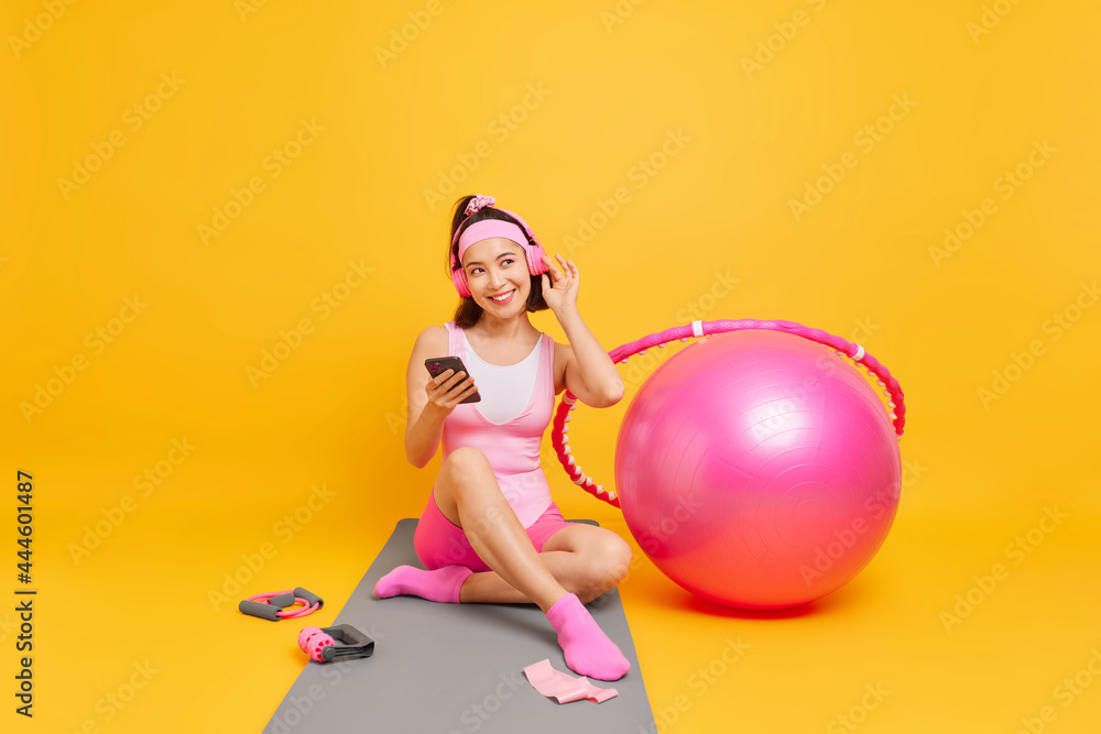 Pleased Asian woman enjoys favorite music from playlist wears wireless headphones holds mobile phone sits on fitness mat goes in for sport rests after domestic workout isolated over yellow wall Pleased Asian woman enjoys favorite music from playlist wears wireless headphones holds mobile phone sits on fitness mat goes in for sport rests after domestic workout isolated over yellow wall