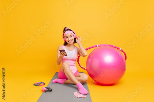 Pleased Asian woman enjoys favorite music from playlist wears wireless headphones holds mobile phone sits on fitness mat goes in for sport rests after domestic workout isolated over yellow wall