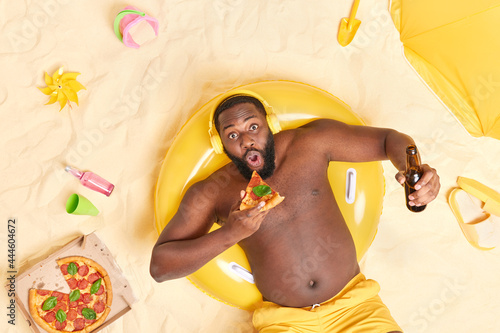 Shocked dark skinned Afro American man eats delicious pizza and drinks beer poses on yellow inflated swimring wears shorts poses at sandy beach listens music via headphones enjoys summer holidays photo