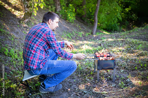 Man in checkered shirt and blue jeans sitting and cooking barbecue in the forest on metal brazier