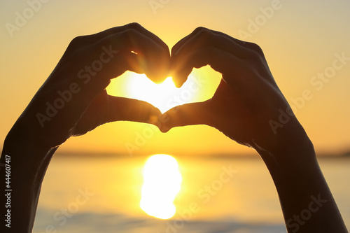 Silhouette of woman's hands in heart shape at sunset against a yellow-orange sky, sunbeams and a sun displayed inside. Symbol of love. Vacation by a sea, ocean, lake, river in summer. Seashore, coast.
