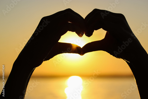 Silhouette of woman s hands in heart shape at sunset against a yellow-orange sky  sunbeams and a sun displayed inside. Symbol of love. Vacation by a sea  ocean  lake  river in summer. Seashore  coast.