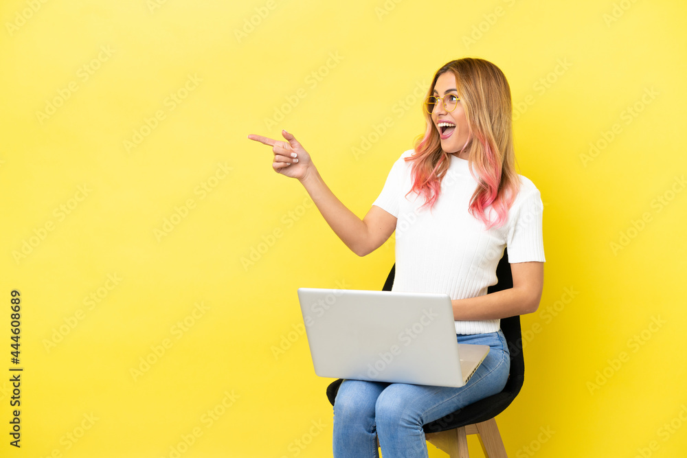Young woman sitting on a chair with laptop over isolated yellow background pointing finger to the side and presenting a product Young woman sitting on a chair with laptop over isolated yellow background pointing finger to the side and presenting a product