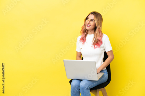 Young woman sitting on a chair with laptop over isolated yellow background thinking an idea while looking up © luismolinero