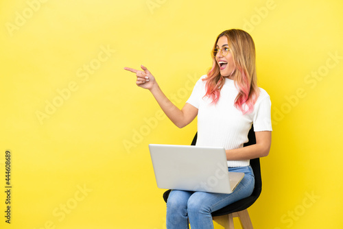 Young woman sitting on a chair with laptop over isolated yellow background pointing finger to the side and presenting a product © luismolinero