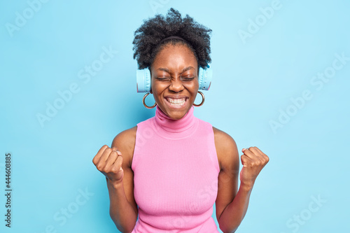 Happy dark skinned woman feels joy of victory shakes fists gestures actively celebrates success clenches teeth listens music via wireless headphones isolated over blue background. Yes I did it