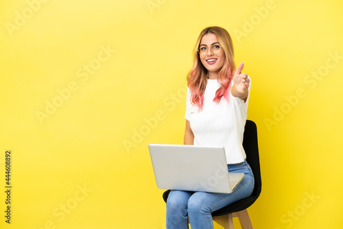 Young woman sitting on a chair with laptop over isolated yellow background shaking hands for closing a good deal © luismolinero