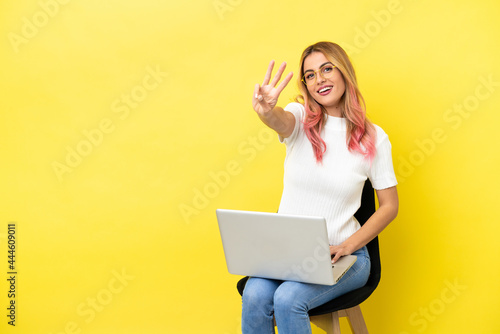 Young woman sitting on a chair with laptop over isolated yellow background happy and counting three with fingers © luismolinero