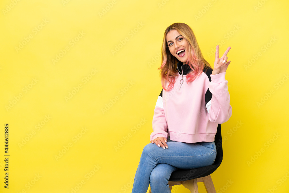 Young woman sitting on a chair over isolated yellow background smiling and showing victory sign Young woman sitting on a chair over isolated yellow background smiling and showing victory sign