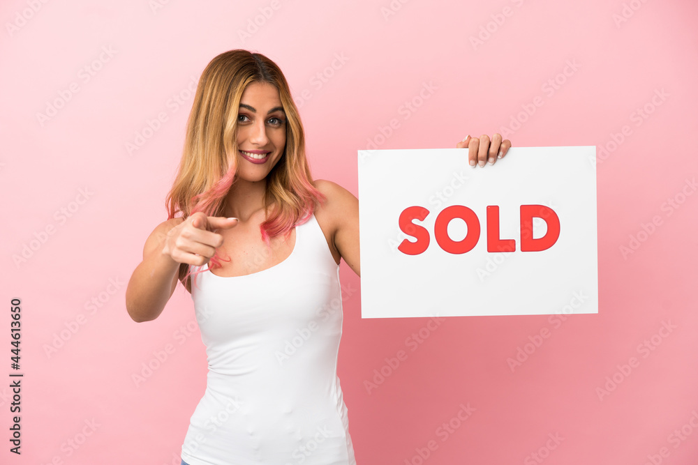 Young woman over isolated pink background holding a placard with text SOLD and pointing to the front Young woman over isolated pink background holding a placard with text SOLD and pointing to the front