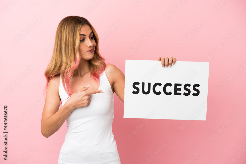 Young woman over isolated pink background holding a placard with text SUCCESS and pointing it Young woman over isolated pink background holding a placard with text SUCCESS and pointing it